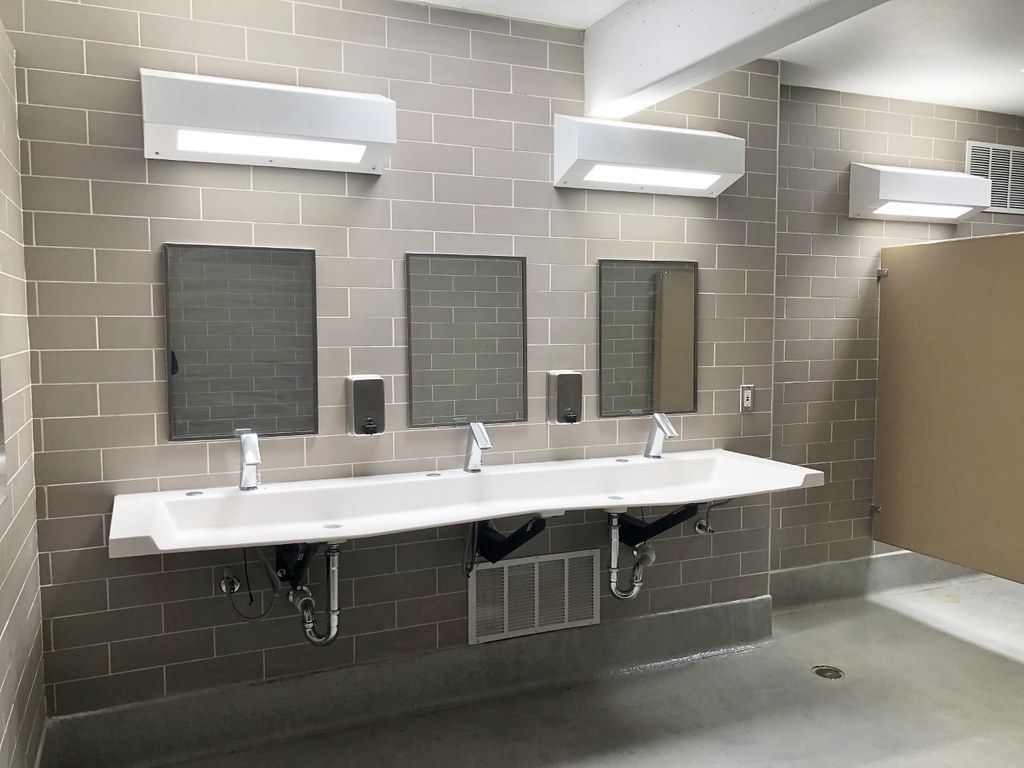 Salle de toilette commerciale : lavabo mural à trois postes, robinets à capteur et plomberie apparente